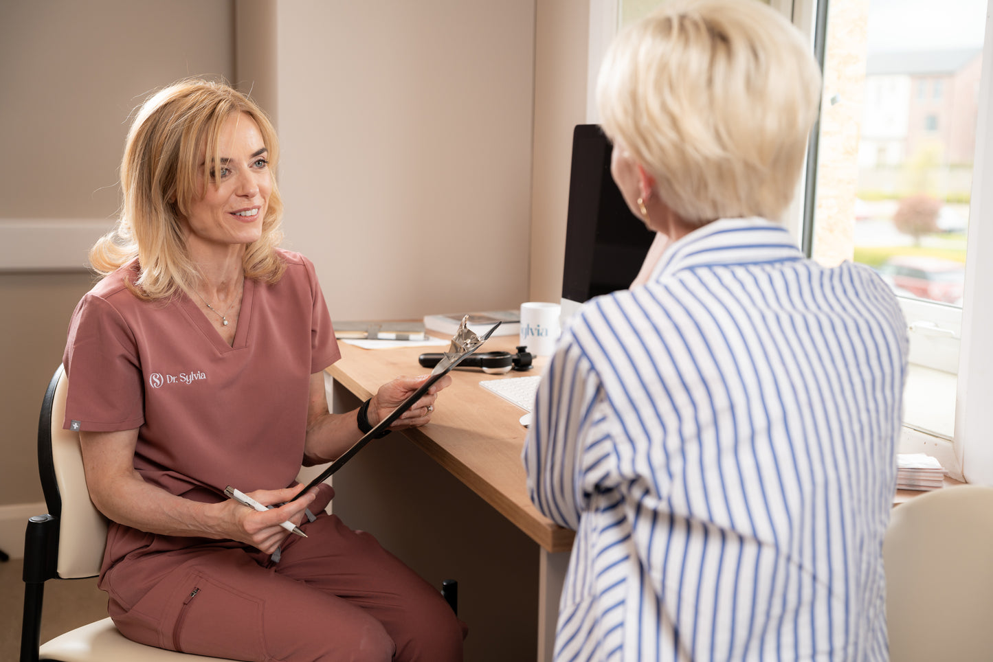 Doctor in pink scrubs sitting with a patient in a medical office setting.