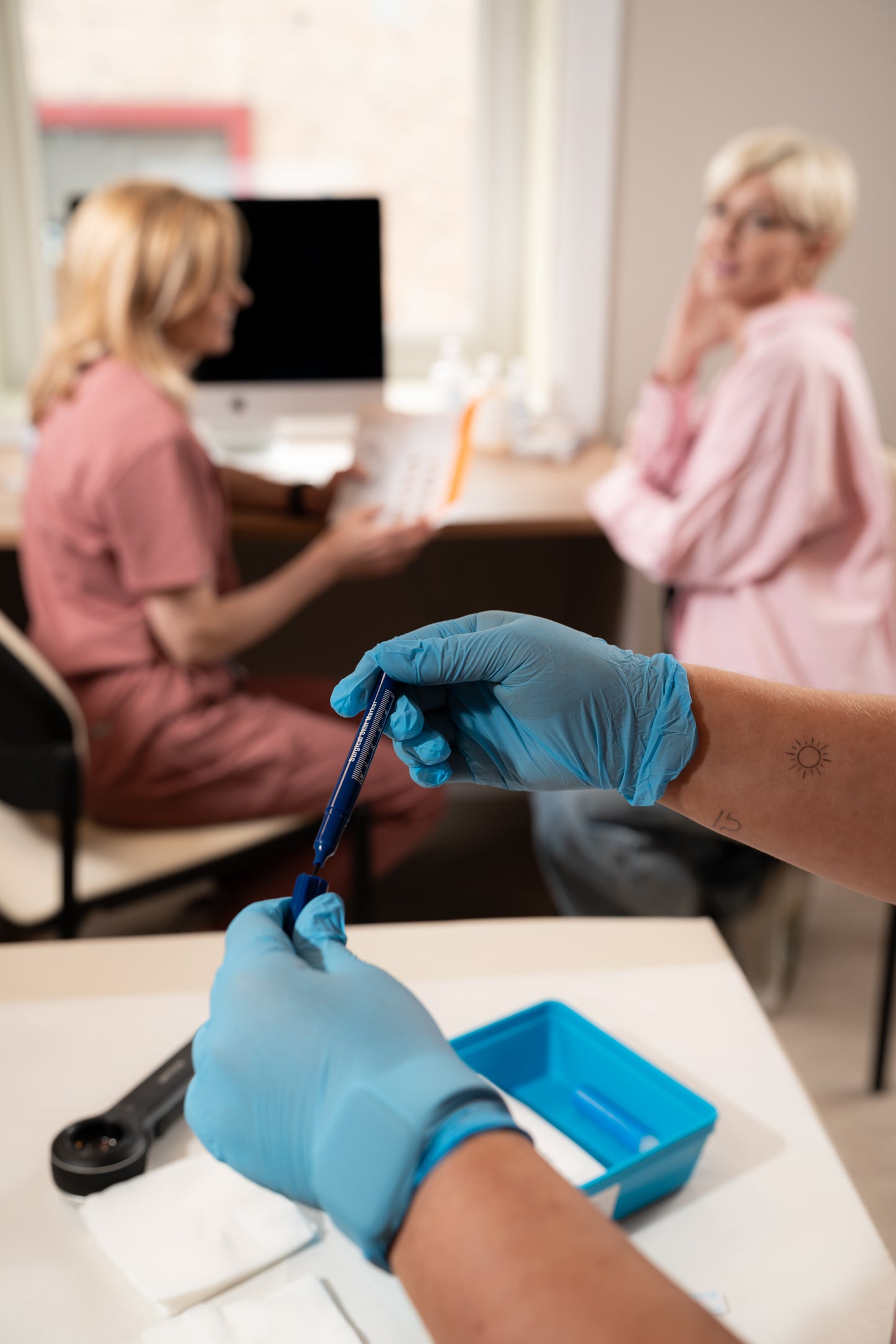 Person wearing blue gloves holding a tool in a medical or clinical setting with another person in the background.